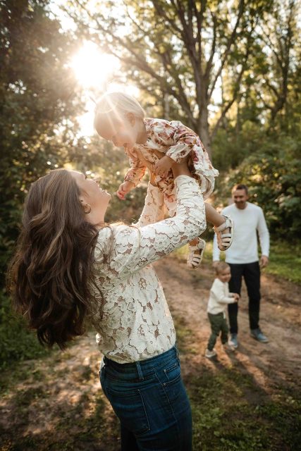 mother playfully throws toddler daughter in floral dress into the air while father and son play in the background.